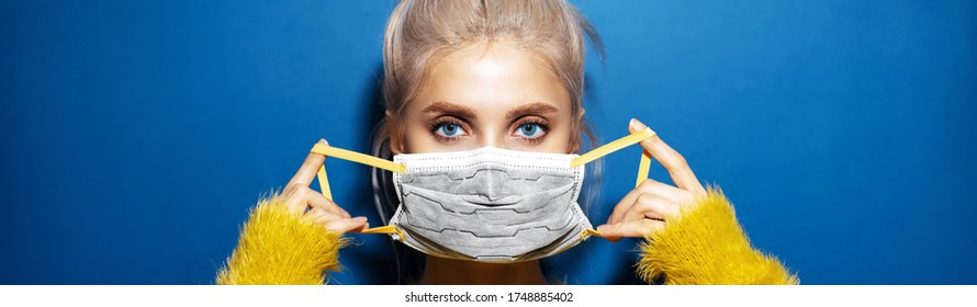 Panoramisch portret van een jong blond meisje met blauwe ogen die een medisch griepmasker op het gezicht zet. Preventie van het coronavirus. Studioachtergrond van blauwe kleur met exemplaarruimte.