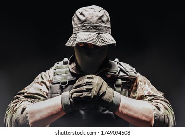 Photo of a fully equipped camouflaged soldier in panama hat posing with fists on dark background wirh fog.