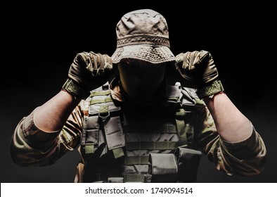 Photo of a fully equipped camouflaged soldier putting on panama hat with tactical vest and gloves  on dark background wirh fog.