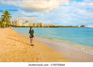 Biracial Asian teenage girl walking alone at empty Ala Moana Beach along the ocean at sunset. Waikiki, Island Colony and Diamond Head visible in background