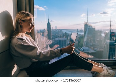 Happy female blogger watching positive video vlog on streams website connecting to 4g internet on laptop computer, cheerful hipster girl networking near panoramic window with metropolitan scenery