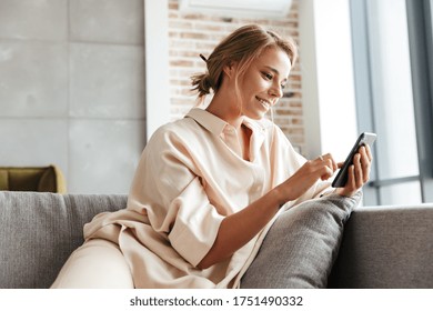 Image of happy nice woman in pajamas smiling and using cellphone while sitting on sofa at living room