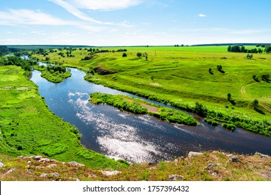 view from the top of the cliff to the river with the island in the middle. blue sky is reflected in the water of the river. hills with alpine meadows