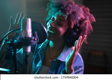 ヘッドフォンを着用した若いプロの笑顔のエネルギッシュなアフリカの女性歌手が、背景にカラフルなライトを備えた音楽スタジオで録音しながら、マイクを使って新しい曲を演奏しています。
