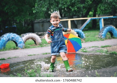 Little cute boy in denim overalls shorts and green rubber boots plays in a large puddle. He launches boats and jumps in a puddle.