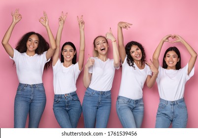 Cinco chicas positivas gesticulando diferentes símbolos posando sobre fondo de estudio rosa, sonriendo a la cámara.