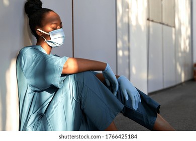 Perawat scrub afrika wanita yang lelah memakai masker wajah, sarung tangan seragam biru duduk di lantai rumah sakit. Depresi sedih dokter etika hitam merasa kelelahan kelelahan stres, kurang tidur, tidur siang di tempat kerja.