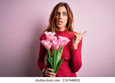 Junge schöne brünette Frau, die einen Strauß rosa Tulpen über isoliertem Hintergrund hält Überrascht, mit dem Finger zur Seite zeigend, offener Mund, erstaunter Ausdruck.