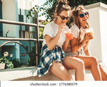 Two young beautiful smiling hipster girls in trendy summer clothes. Carefree women posing outdoors.Positive models holding and drinking fresh cocktail smoothie drink in plastic cup with straw