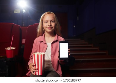 Retrato de vista frontal de una adolescente rubia que muestra un teléfono inteligente con pantalla en blanco y mira la cámara mientras sostiene una taza de palomitas de maíz en el cine, copia espacio