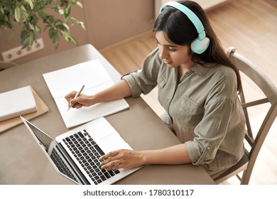 Indian girl student wear headphones learning online watching webinar class looking at laptop computer elearning remote lesson making notes or video calling virtual conference meeting teacher at home.
