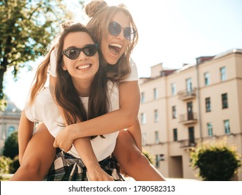 トレンディな夏の白い t シャツの服で 2 つの若い美しい笑顔流行に敏感な女性。通りの背景にポーズをとってセクシーな屈託のない女性。友人の背中に飛び乗り、屋外でおんぶをするモデル