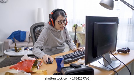 熱意を持ってコンピューター ゲームをプレイ フードで面白いアジア中国の女の子。暇な時間の娯楽のコンセプトの無駄。怠惰な女性は家にいて、オンラインゲームを楽しみ、散らかった寝室でジャンクフードを食べます。