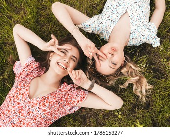 Two young beautiful smiling hipster girls in trendy summer sundress.Sexy carefree women lying on the green grass in sunglasses.Positive models having fun.Top view.They show peace sign