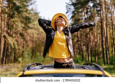 Mujer feliz de pie fuera del techo corredizo del coche amarillo. Niña sonriente estirándose disfrutando de la vista frontal del sol. Hermosa modelo caucásica con chaqueta de cuero, gafas de sol y sombrero en el techo automático