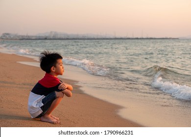 Cute toddler Asian boy sitting alone on the beach at sunset. Child sitting on the beach near sea and looking on the waves, silhouette of the boy on the seaside in sunset.