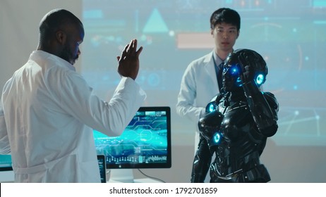 Interracial research team testing the new futuristic automated android in lab. Cybernetic black robot waving hand repeating scientist moves. Artificial intelligence.