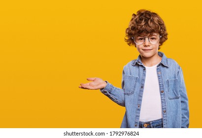 Niño lindo en camisa de mezclilla y gafas mirando a cámara y apuntando al espacio en blanco aislado sobre fondo amarillo