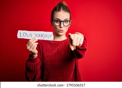 Young beautiful redhead woman holding paper with love yourself message pointing with finger to the camera and to you, hand sign, positive and confident gesture from the front