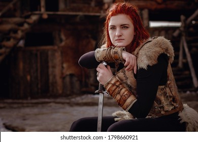 Chica vikinga de pelo rojo. En manos de la espada para luchar contra el enemigo. Reconstrucción de una escena medieval. En el contexto de un gran pueblo vikingo.