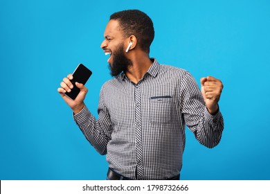 Music Lover. Portrait of overjoyed black male model singing and using his smart phone as mic on blue studio wall