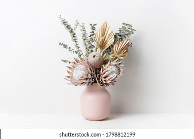 Lindo arranjo de flores secas em um elegante vaso rosa. O buquê inclui proteas-rei rosa, banksias, folhas de eucalipto e ramos de palmeira dourados, fotografados sobre um fundo branco.