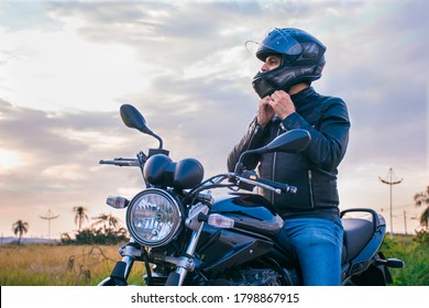 Man sitting on a motorcycle, wearing jeans and a black jacket, fastening his helmet with a landscape in the background.
