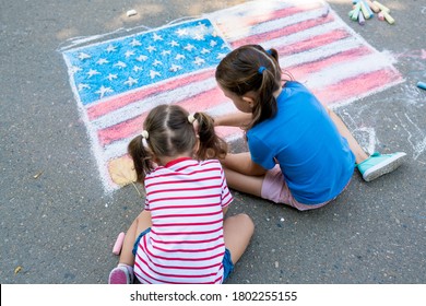 Dua gadis manis menggambar bendera Amerika dengan kapur berwarna di trotoar dekat rumah pada hari musim panas yang cerah. Anak-anak melukis di luar. Perkembangan kreatif anak-anak. Konsep hari patriotik