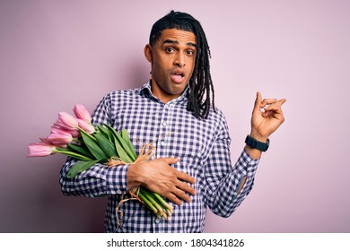 Junger afrikanisch-amerikanischer afroromantischer Mann mit Dreadlocks, der einen Strauß rosa Tulpen hält Überrascht, mit dem Finger zur Seite zeigend, offener Mund, erstaunter Ausdruck.