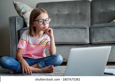 Serious cute little girl 8 years old in a striped T-shirt and jeans with glasses sits at home on a carpet in front of a laptop, remote education technologies and homework