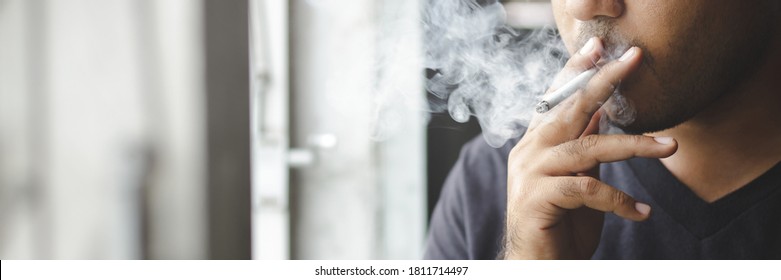Asiatischer Mann, der eine Zigarette raucht, steht am Fenster, dunkler Hintergrund. Konzept Gesundheitswesen. Platz für Bannerkopien.