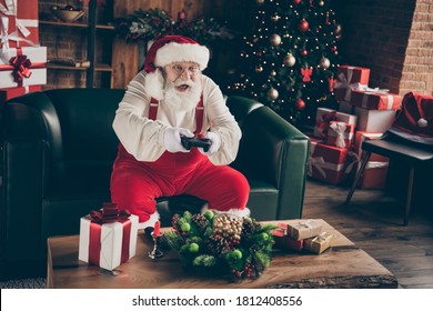 Foto van opgewonden verslaafd grijs haar kerstman zitten bank vieren kerstmis kerstmis nacht middernacht spelen videogame dragen rode pet hoofddeksels in huis binnenshuis nieuwjaar ornament