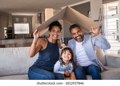 ソファに座って段ボールの屋根を持ち、カメラを見ている笑顔の家族のポートレート。新しい家でソファに座っている間、頭の上に段ボールの屋根を保持している娘を持つアフリカとインドの両親