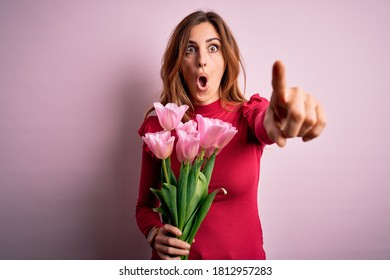 Junge schöne brünette Frau, die einen Strauß rosafarbener Tulpen über isoliertem Hintergrund hält und mit dem Finger überrascht nach vorne zeigt, offener Mund überraschter Ausdruck, etwas auf der Vorderseite