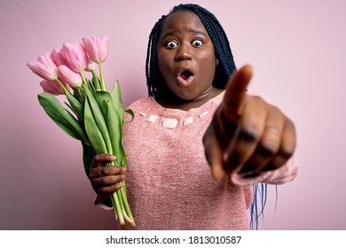 Junge Afroamerikanerin in Übergröße mit Zöpfen, die einen Blumenstrauß aus rosafarbenen Tulpen halten. Zeigen mit dem Finger überrascht nach vorne, offener Mund überraschter Ausdruck, etwas auf der Vorderseite