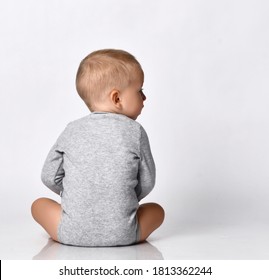 Toddler one-year-old baby boy in diaper and grey one-piece bodysuit with long sleeves sits back to camera looking aside with interest reading. Happy infancy and babyhood concept