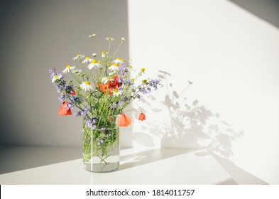Strauß Wildblumen in einer kleinen Glasvase auf dem weißen Tisch. Mohnblumen, Kamille, Kornblumen, grünes Gras. Sommerfoto. Kontrastschatten an der weißen Wand. Landhausstil.