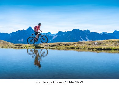 Una mujer agradable montando su bicicleta de montaña eléctrica en los Dolomitas de los Tres Picos, reflejándose en el agua azul de un lago frío de montaña