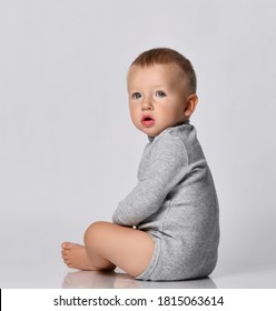 Toddler one-year-old baby boy in diaper and grey one-piece bodysuit with long sleeves sits side and looking to camera with interest. Happy infancy and babyhood concept
