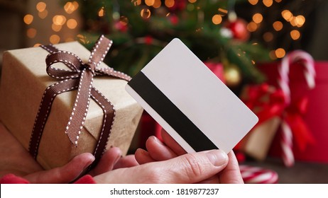 Wanita muda memegang kartu kredit dan kotak hadiah dengan latar belakang dekorasi dan hadiah Natal, close-up. Belanja Natal dan Tahun Baru di Internet, pembayaran dengan kartu kredit.