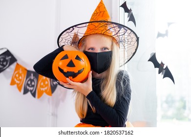Retrato de niña rubia en traje de bruja con máscaras faciales con cubo de dulces en la pandemia de coronavirus preparándose para Halloween protegiéndose de COVID-19.