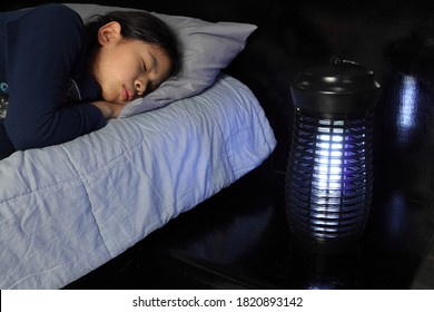 虫や蚊の電気殺虫器の近くで眠っているかわいい若い女の子。暗い寝室の木製のベッドテーブルに青いライトランプが置かれ、虫から夜間の安全な睡眠環境を確保しています。