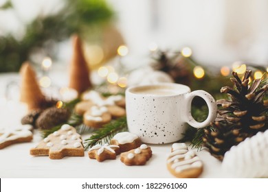 Weihnachtslebkuchenplätzchen, Kaffee in stilvoller weißer Tasse, Tannenzapfen und warme Lichter auf weißem Holztisch. Hallo Winter, gemütliches, stimmungsvolles Bild mit selektivem Fokus