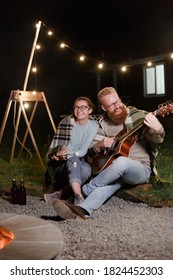Young couple, caucasian woman and red haired bearded man, on a night picnic with a guitar and marshmello have a good time