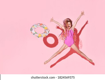Fille heureuse avec des tresses colorées en maillot de bain une pièce rose, sautant et criant de joie, lançant une bouée en caoutchouc avec un motif de fruits. Plan complet isolé sur rose. Enfance, émotions, été
