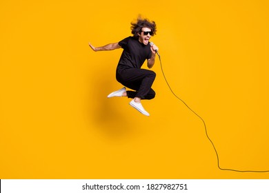 Volledige lengte lichaamsgrootte portret van springende man met zwarte kleding die luid zingt in microfoon geïsoleerd op felgele kleur achtergrond