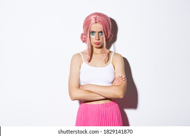 Portrait of angry cute girl with pink wig and glamour makeup, cross arms on chest and sulking mad at someone, feeling offended, standing over white background