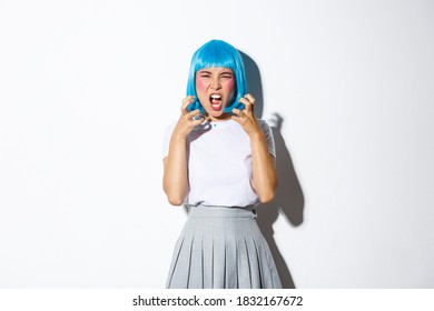 Portrait of angry asian girl in blue wig, yelling at someone with hatred, standing over white background