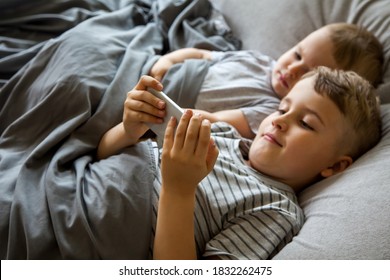 Twee kleine jongens met bruin haar liggen in een groot bed. Lach, kijk naar tekenfilms op de telefoon en tablet. Speel de gadget. Grijs beddengoed. Kinderen zijn blank.
