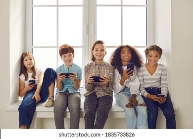 Beragam kelompok anak-anak berusia 9-10 tahun yang bahagia bermain game online di ponsel. Anak laki-laki dan perempuan yang tersenyum memegang perangkat seluler dan melihat kamera duduk di ambang jendela dengan jendela mockup putih